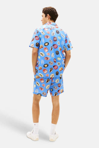 Blauw overhemd met korte mouwen van Tommy Jeans, voorzien van retro badge-patroon en gedragen met bijpassende shorts.
