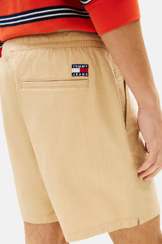 Lichtbruine short van TOMMY JEANS met elastische tailleband, een paspelzak en een kleine merkpatch op de rug. Gedragen door een man met een rood gestreept shirt.