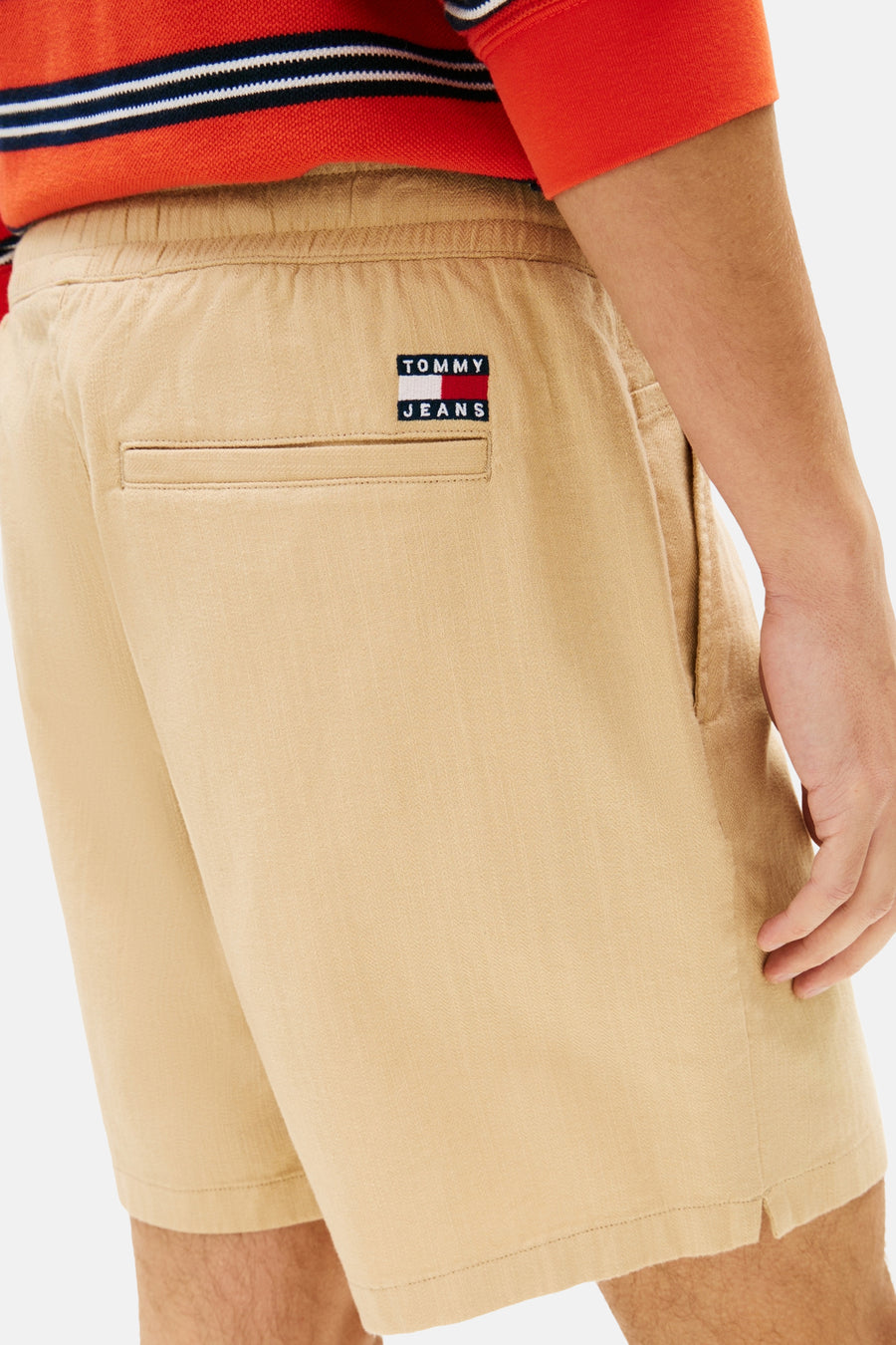 Lichtbruine short van TOMMY JEANS met elastische tailleband, een paspelzak en een kleine merkpatch op de rug. Gedragen door een man met een rood gestreept shirt.
