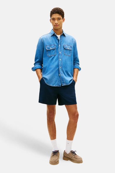 Blauwe short van TOMMY JEANS, gecombineerd met een denim overhemd en bruine boots met witte sokken.
