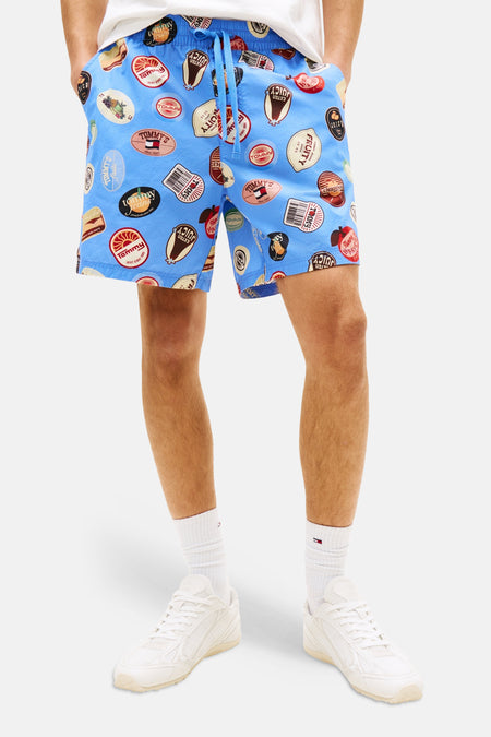 Blauwe short van TOMMY JEANS, bedrukt met diverse 'Tommy's'-fruitstickers, gedragen met witte sneakers en sokken.
