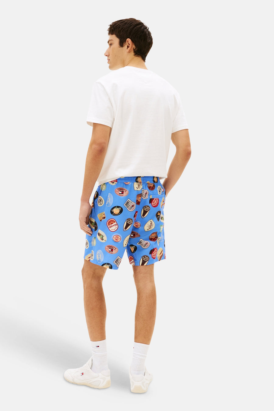Blauwe short van TOMMY JEANS met een opvallende badgeprint, gecombineerd met een wit T-shirt en witte sokken met sneakers.
