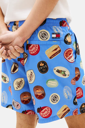 Blauwe short van TOMMY JEANS met vintage fruitstickers, achterzak en zichtbare tailleband, gedragen met een wit T-shirt.
