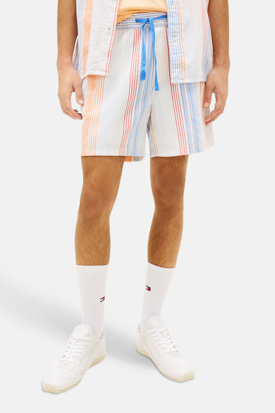 Multicolor short van TOMMY JEANS met verticale oranje, rode en blauwe strepen, blauw trekkoord, witte sokken en sneakers.