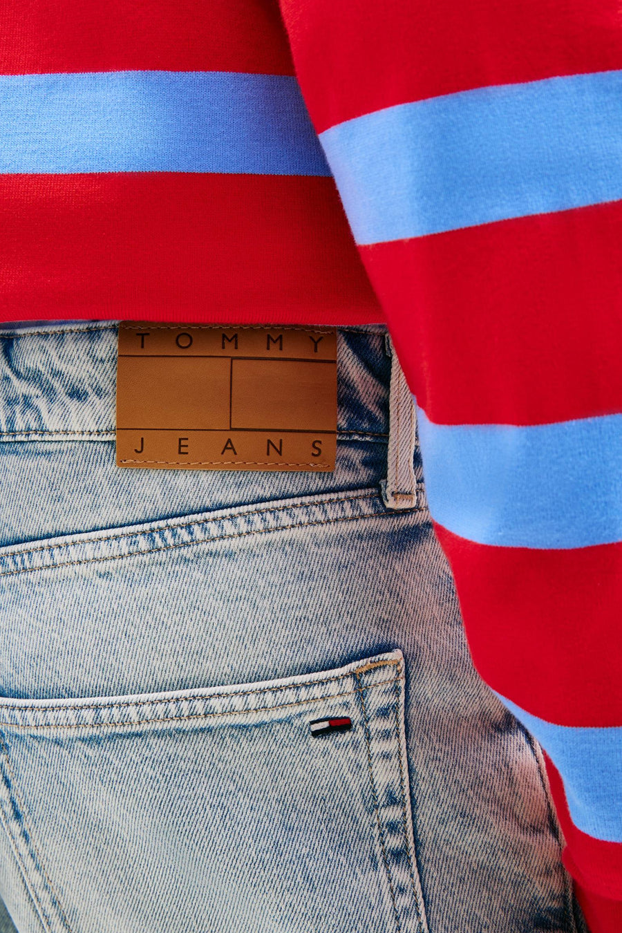 Short en jean denim bleu clair de TOMMY JEANS avec patch en cuir marron et petit logo drapeau, porté avec un pull à rayures rouge et bleu.