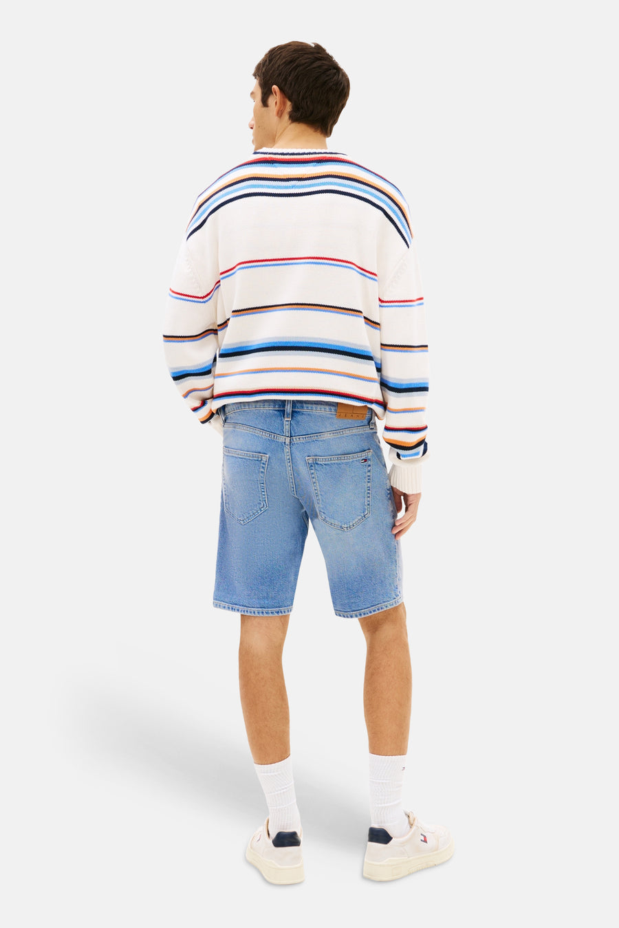 Mid-blauwe denim jeansshort van TOMMY JEANS met achterzakken en bruin embleem, gedragen door een man in een gestreepte trui.