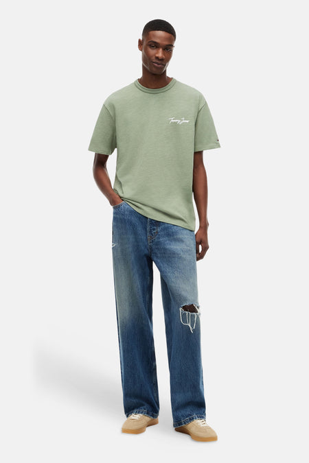T-shirt vert à manches courtes TOMMY JEANS avec logo script blanc sur la poitrine, porté par un homme avec un jean déchiré et des baskets beiges.
