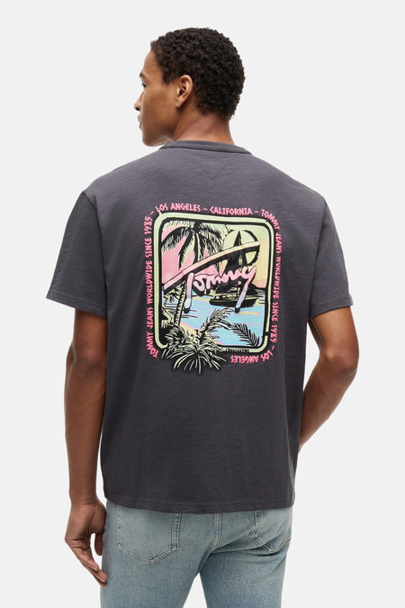 Grijs T-shirt met korte mouwen van TOMMY JEANS, achterkant met kleurrijke retro strandprint en tekst, gedragen met jeans.