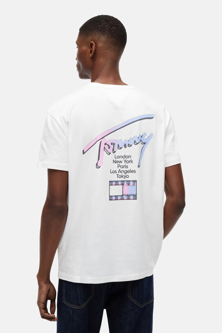 T-shirt blanc à manches courtes TOMMY JEANS, avec logo script dégradé rose-bleu, liste de villes et logo drapeau au dos, porté avec un jean foncé.