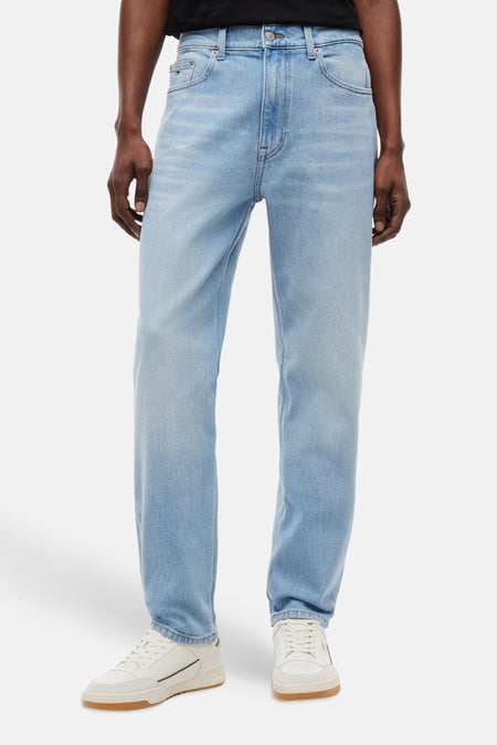 Jean fuselé bleu clair de TOMMY JEANS, porté par un homme avec un t-shirt noir et des baskets blanches.