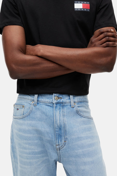 Jean slim fuselé bleu clair en denim de TOMMY JEANS, avec fermeture à boutons, poches avant et un petit logo drapeau sur la poche ticket, porté avec un t-shirt noir.