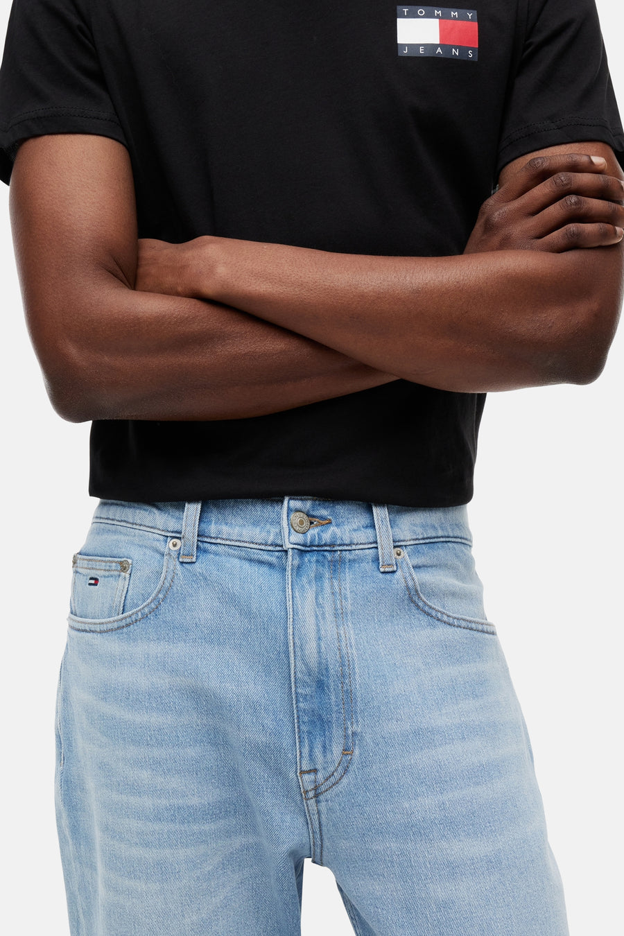 Jean slim fuselé bleu clair en denim de TOMMY JEANS, avec fermeture à boutons, poches avant et un petit logo drapeau sur la poche ticket, porté avec un t-shirt noir.