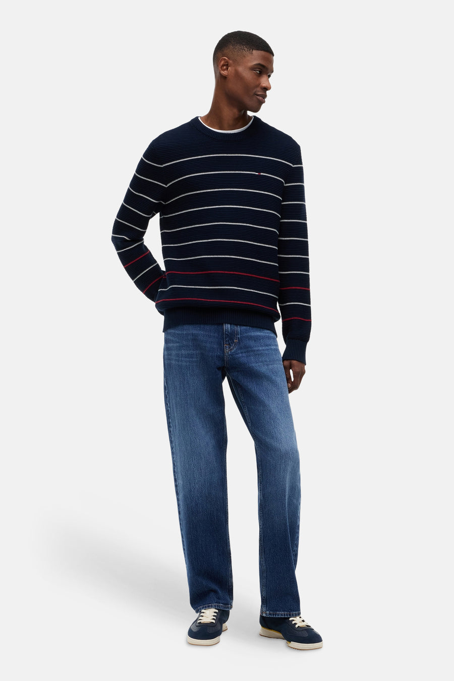 Blauwe trui met ronde hals van TOMMY JEANS, met witte en rode strepen, ribgebreid, gedragen door een man met jeans en sneakers.