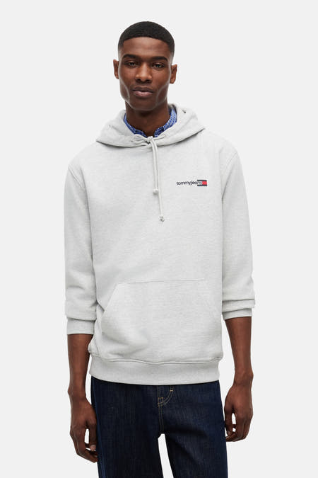 Grijze sweater van TOMMY JEANS in hoodie-stijl met klein borstlogo, gedragen over een blauw geruit overhemd en donkere jeans.