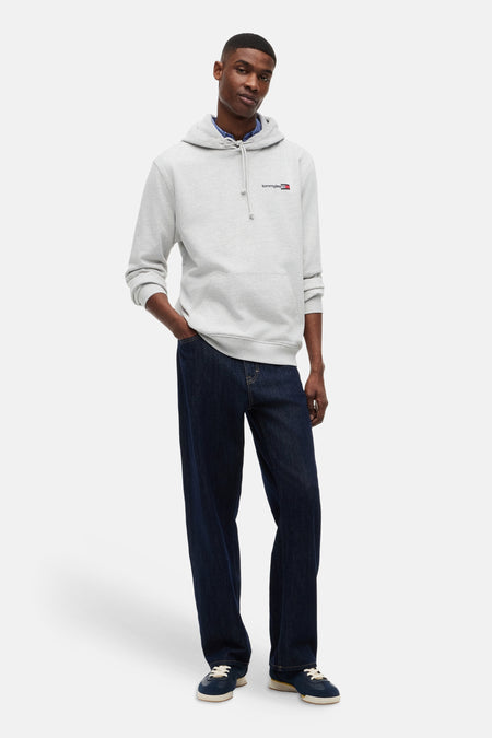 Grijze sweater van TOMMY JEANS met capuchon en logo op de borst, gedragen over een blauw overhemd met donkerblauwe jeans en blauwe sneakers.
