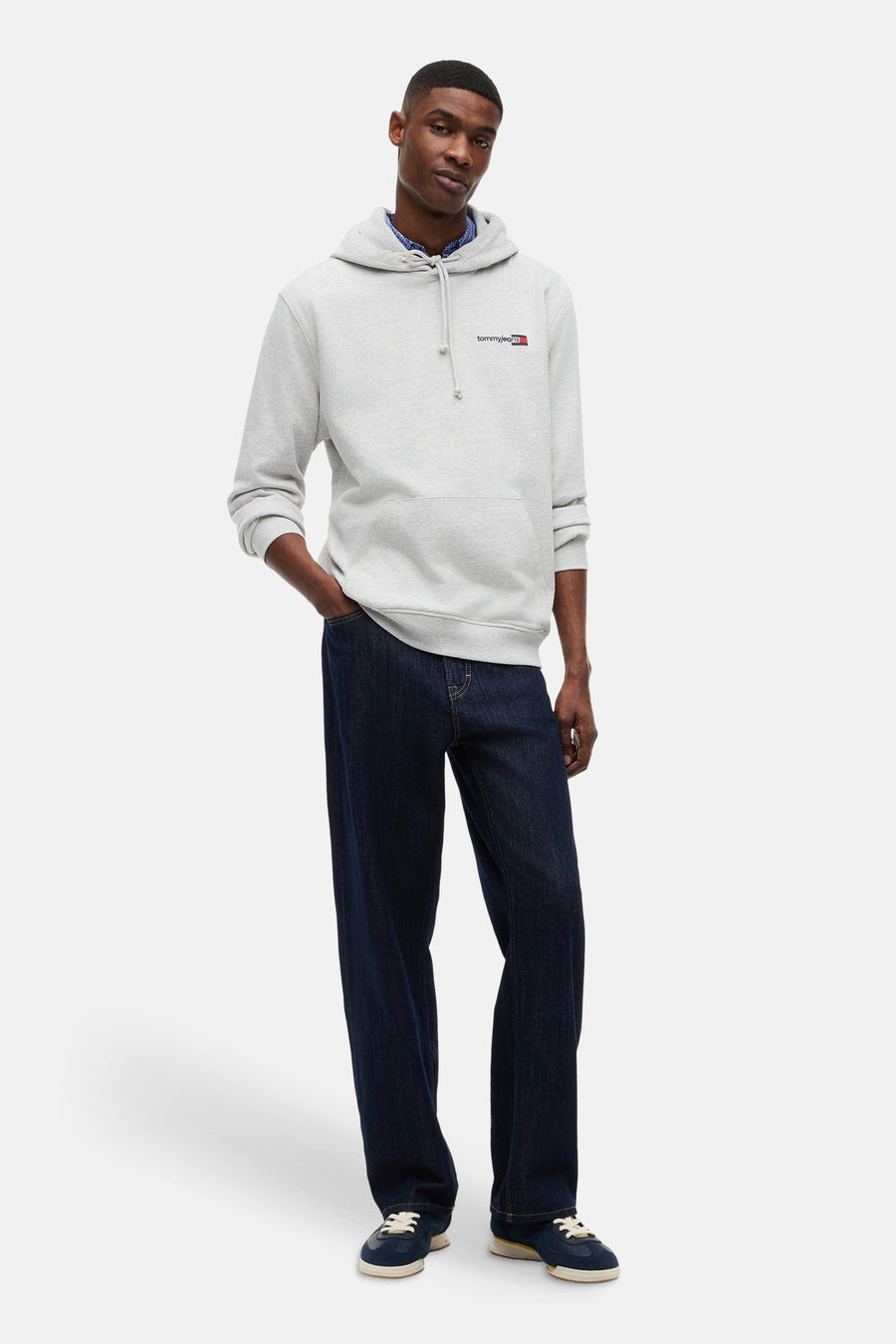 Grijze sweater van TOMMY JEANS met capuchon en logo op de borst, gedragen over een blauw overhemd met donkerblauwe jeans en blauwe sneakers.