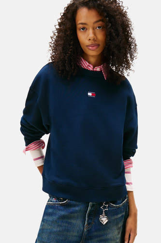Blauwe sweater van TOMMY JEANS, gedragen over een gestreept overhemd en een spijkerbroek met een bedel.
