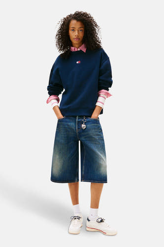 Blauwe sweater van TOMMY JEANS, gedragen over een gestreept overhemd, met wijde denim bermuda en witte sneakers.
