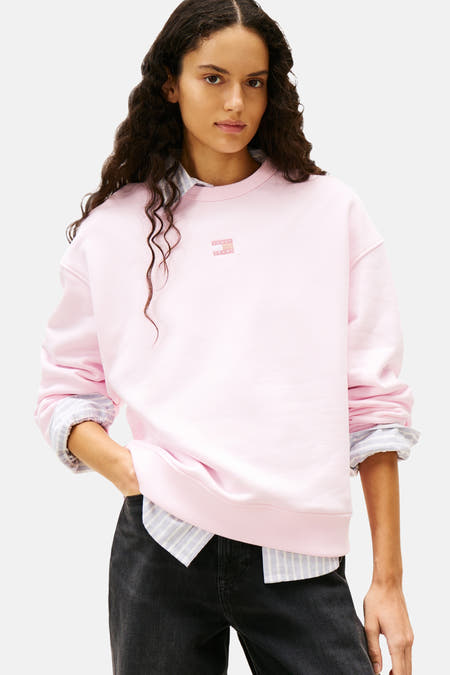 Pull-over rose de TOMMY JEANS, avec des manches longues et un col rond, porté avec une chemise rayée et des jeans noirs.