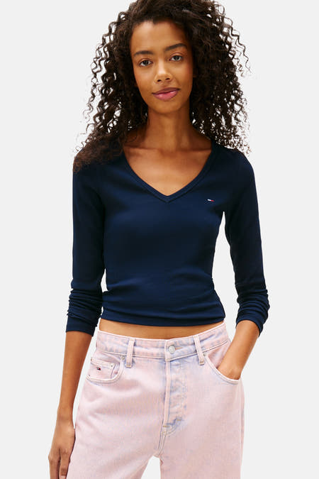 T-shirt bleu à manches longues de TOMMY JEANS, avec col V, associé à des jeans roses et un pull autour de la taille.