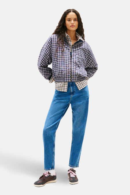 Vrouw in geruite jas, jeans en sneakers, staand.
