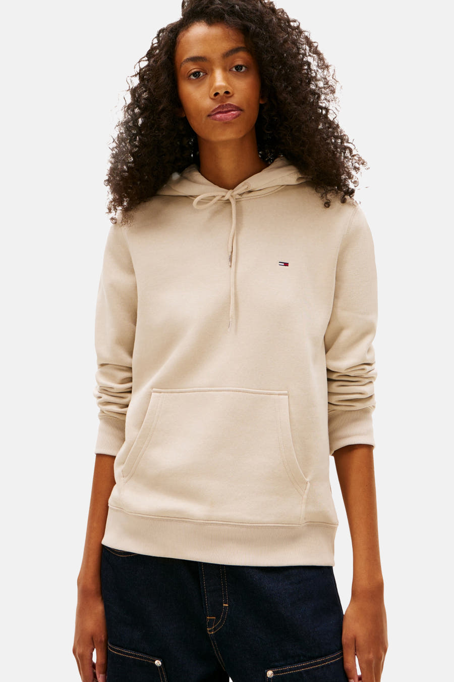 Beige sweater van TOMMY JEANS met capuchon en trekkoord, gedragen met donkerblauwe jeans.
