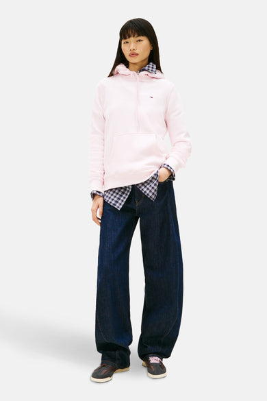 Roze sweater van TOMMY JEANS, met een capuchon, gedragen over een blauw geruit overhemd en wijde, donkerblauwe jeans.
