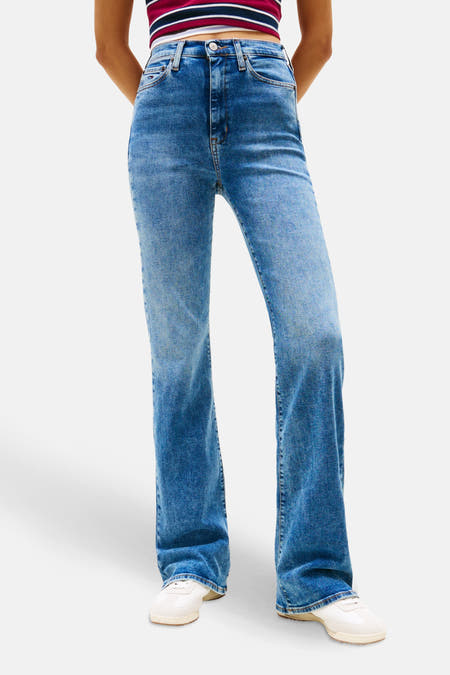 Jean flare en denim bleu moyen de TOMMY JEANS, avec une jambe qui s'élargit et porté avec un chemisier rayé et une veste bomber marine.