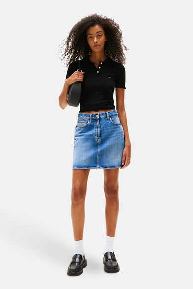 Polo noir à manches courtes de TOMMY JEANS, avec des boutons blancs, associé à une jupe en denim, des chaussettes blanches et des chaussures noires.