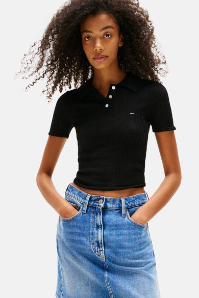Polo noir à manches courtes de TOMMY JEANS, avec fermeture à boutons et porté avec une jupe denim bleue.