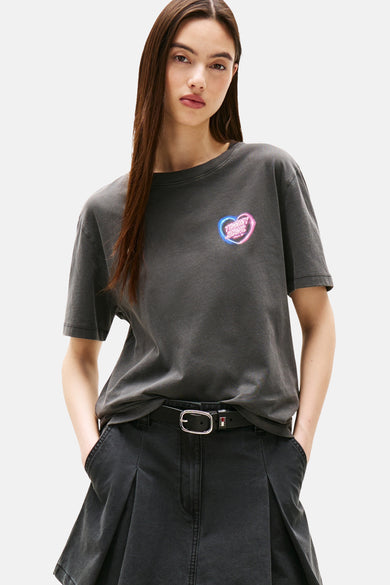 Grijs T-shirt met korte mouwen van TOMMY JEANS, met een neon hartvormig logo op de borst en gedragen met een zwarte broek en riem.
