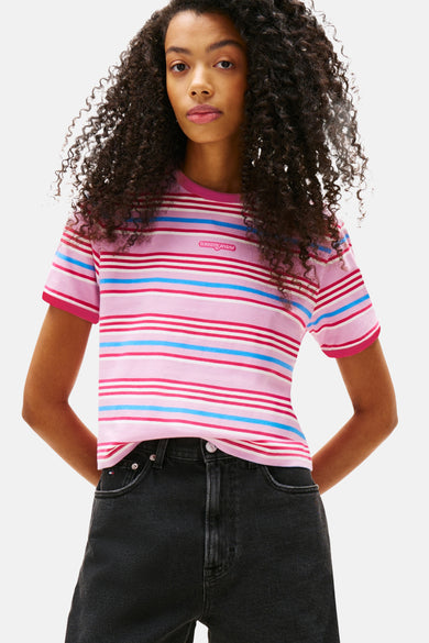 T-shirt rose à manches courtes de TOMMY JEANS, avec un motif rayé en rose, blanc et bleu, porté avec des jeans noirs.