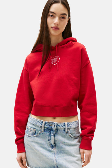 Pull-over rouge de TOMMY JEANS avec un logo en forme de cœur blanc, porté avec un jean à coupe basse.