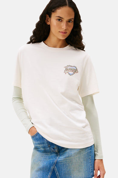 T-shirt blanc à manches courtes de TOMMY JEANS, avec imprimé logo sur la poitrine et associé à un t-shirt à manches longues et une jupe en denim.