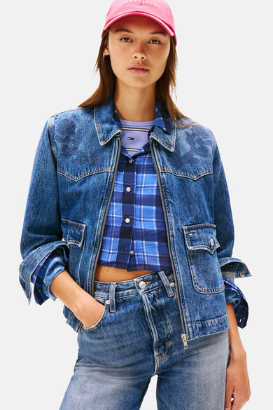Denim jeansjas in middenblauw denim van Tommy Jeans, met bloemenborduursel op de schouders, gecombineerd met blauw geruit overhemd en roze pet.
