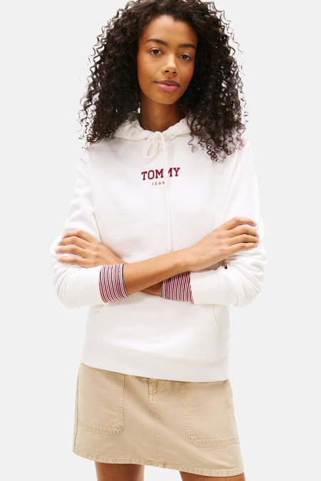 Pull-over blanc à capuche de TOMMY JEANS, avec des manches rayées et associé à une jupe beige, des chaussettes blanches et des baskets crème.