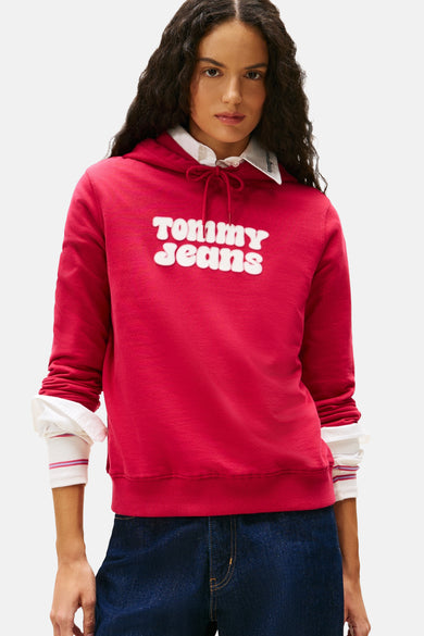 Pull-over rose Tommy Jeans avec capuche, manches longues et logo blanc imprimé sur l'avant, porté sur un chemisier blanc.