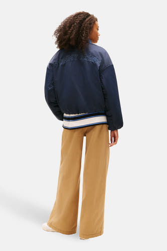 Blauwe korte jas van TOMMY JEANS, met 'Tommy Jeans' op de rug en beige broek.
