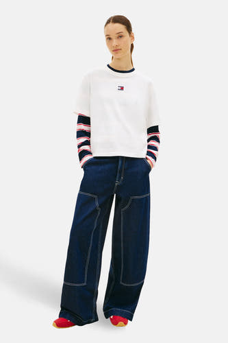 Wit T-shirt met korte mouwen van TOMMY JEANS, gedragen over een gestreept shirt met wijde donkere jeans en rode schoenen.
