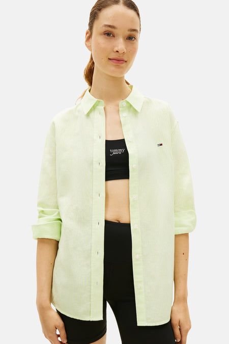Chemise vert clair à manches longues avec de fines rayures verticales de TOMMY JEANS, portée ouverte sur un soutien-gorge de sport noir et un short cycliste noir.