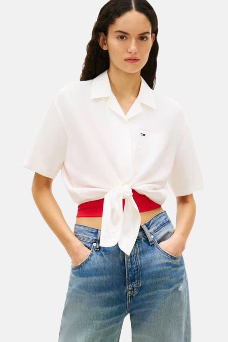 Wit overhemd met korte mouwen van TOMMY JEANS, geknoopt in de taille, met rode top en blauwe jeans.
