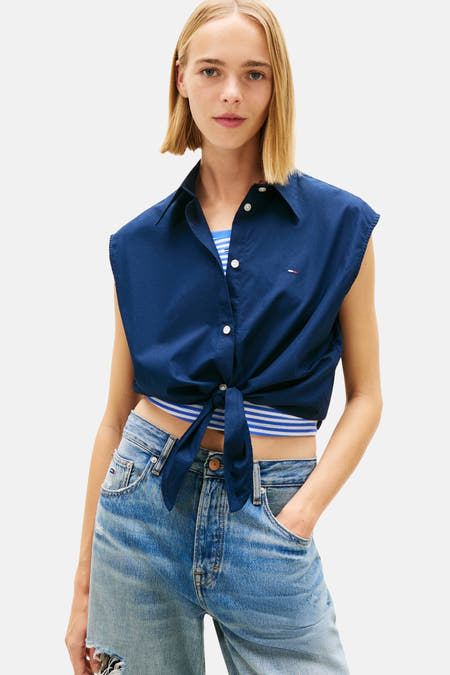 Chemisier bleu sans manches de Tommy Jeans, noué sur le devant, avec un jean bleu clair à jambes larges et des chaussures blanches.