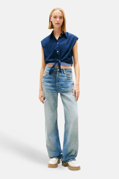 Mouwloze blauwe blouse van Tommy Jeans, geknoopt aan de voorkant, met een lichtblauwe jeans met wijde pijpen en witte schoenen.
