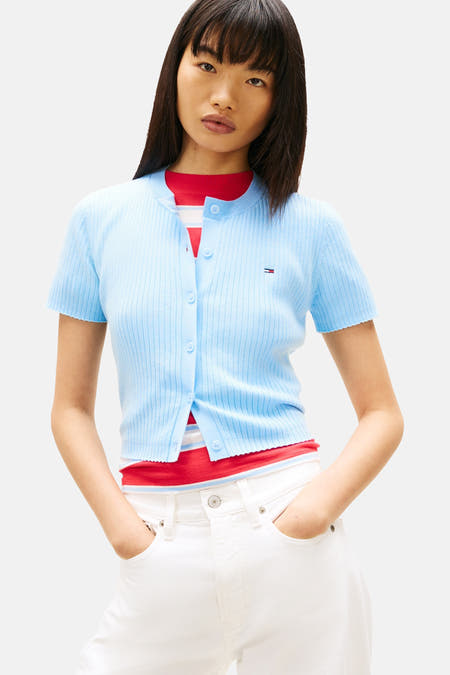 Gilet bleu de TOMMY JEANS, porté sur une chemise rouge et associé à un pantalon blanc et un sac à main marron.