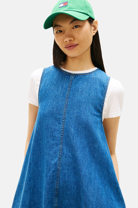 Mouwloos, midblauw denim kleedje van Tommy Jeans, gecombineerd met een wit T-shirt en groene pet.