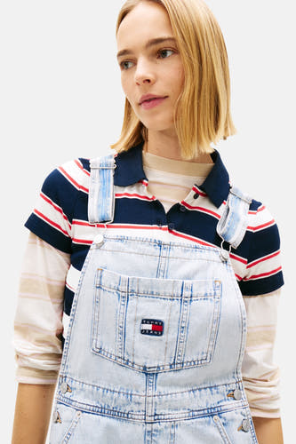 Lichtblauwe denim jumpsuit van TOMMY JEANS, gedragen over een gestreept poloshirt en gestreept T-shirt met lange mouwen, met logo patch op de borst.