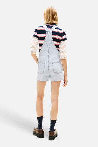 Lichtblauwe denim jumpsuit van TOMMY JEANS, achterkant zichtbaar, gedragen over een gestreept shirt, met marineblauwe sokken en bruine loafers.
