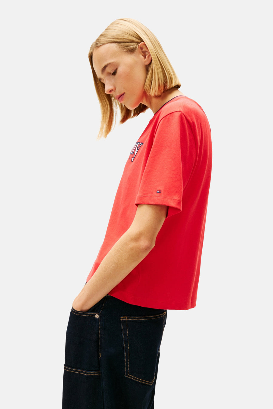 Rood T-shirt met korte mouwen van TOMMY JEANS, met een logo op de borst en donkere jeans met zichtbare stiksels.

