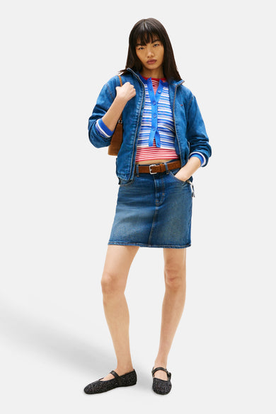 Korte donkerblauwe denim rok van TOMMY JEANS, gecombineerd met een bruine riem, een gestreepte top en een spijkerjasje.
