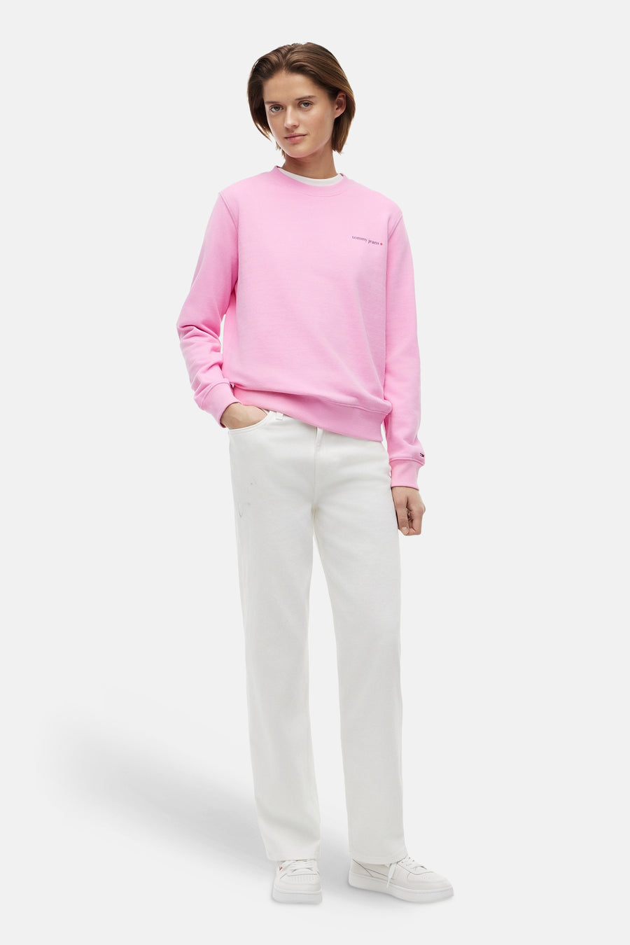 Pull rose à col rond de TOMMY JEANS, avec le texte 'tommy jeans' sur la poitrine, logo drapeau sur le poignet, et stylisé avec un jean blanc et des baskets.